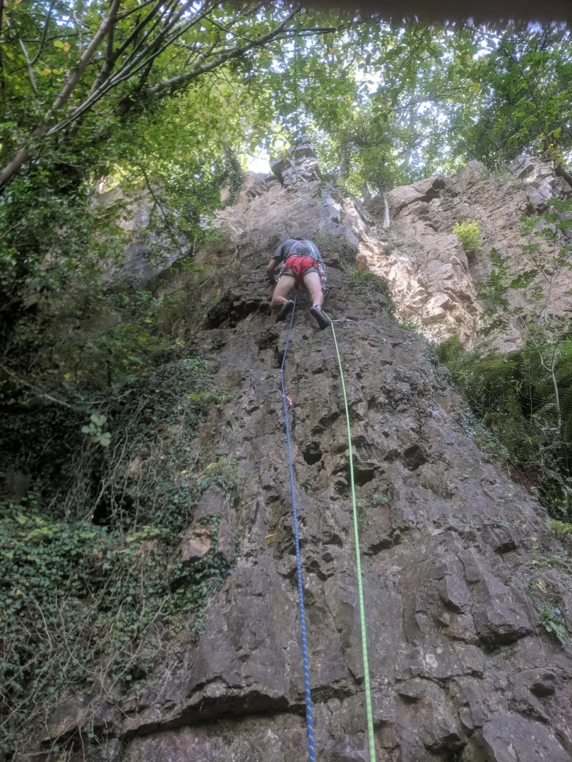 A man in red shorts climbs the vertical limestone crag at Symonds Yat, seen from below with brightly coloured ropes leading down to the camera