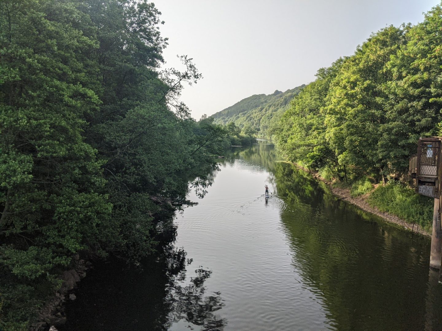 A paddle boarder moves upstream in the mid-distance, on the Rive Wye. Thickly wooded hills rise steeply from the banks.